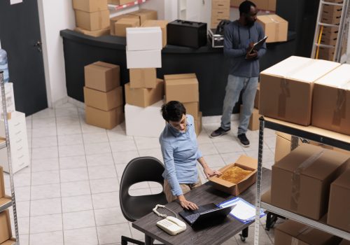 Top view of warehouse supervisor packing client clothes order in cardboard box while checking shipping details on laptop computer. Team working in storehouse in delivery department.